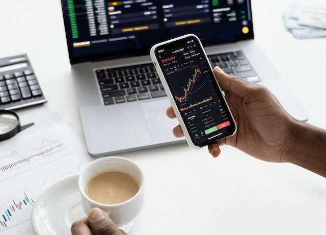 Man looking at stock trends on his smartphone trading app while checking stock trends also on his laptop